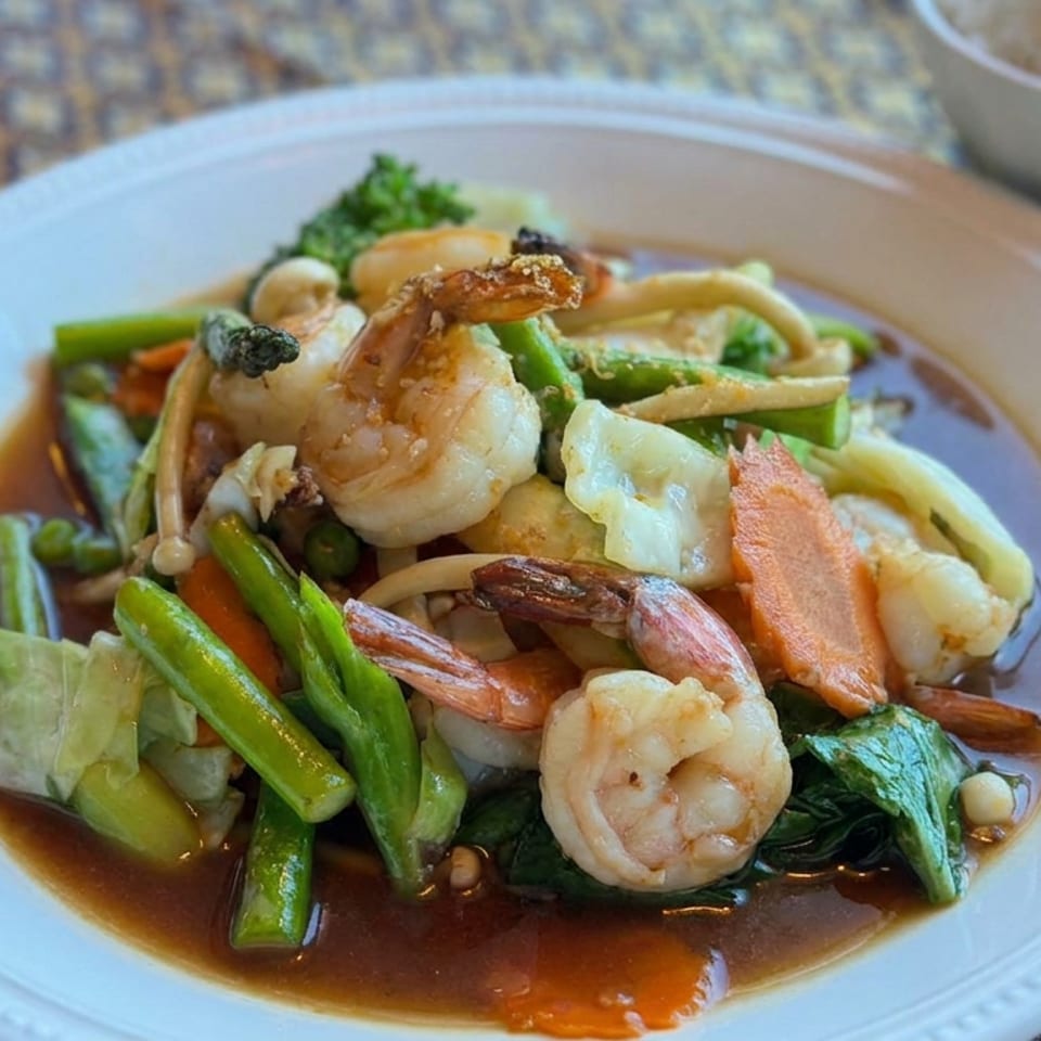 Thai-style stir-fried shrimp with mixed vegetables in savory sauce, served on a white plate beside a bowl of rice.
